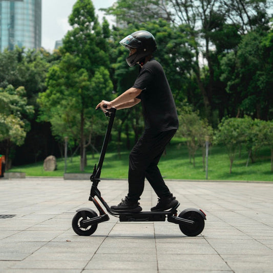 Person riding an Apollo Explore 2.0 electric scooter through an urban park, wearing a helmet and black clothing, with trees and city buildings in the background
